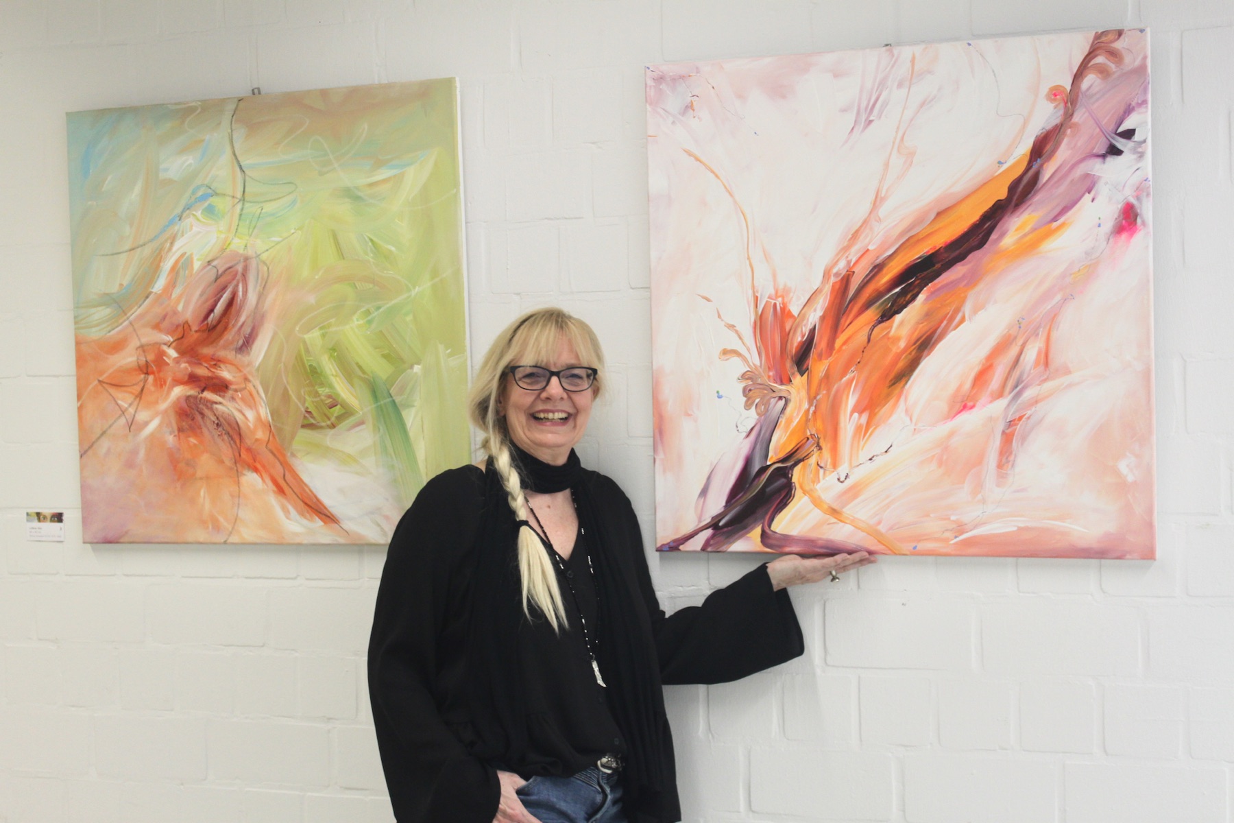 Silvia Grasser lächelt zwischen zwei hellen, zarten Acrylbildern in Pastell und Orange