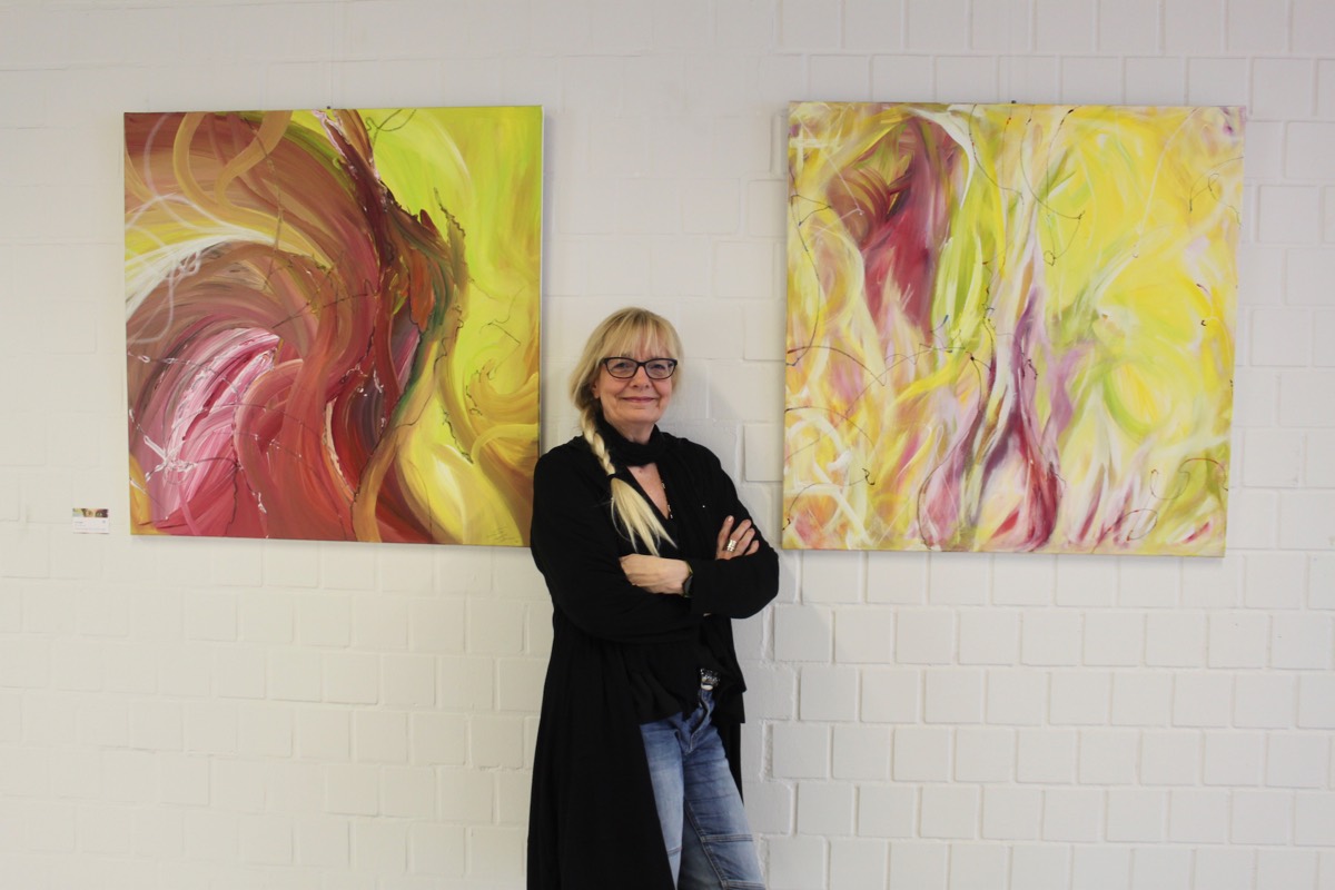 Silvia Grasser steht lächelnd mit verschränkten Armen zwischen zwei quadratischen Acrylbildern in Gelb, Rot und Pink