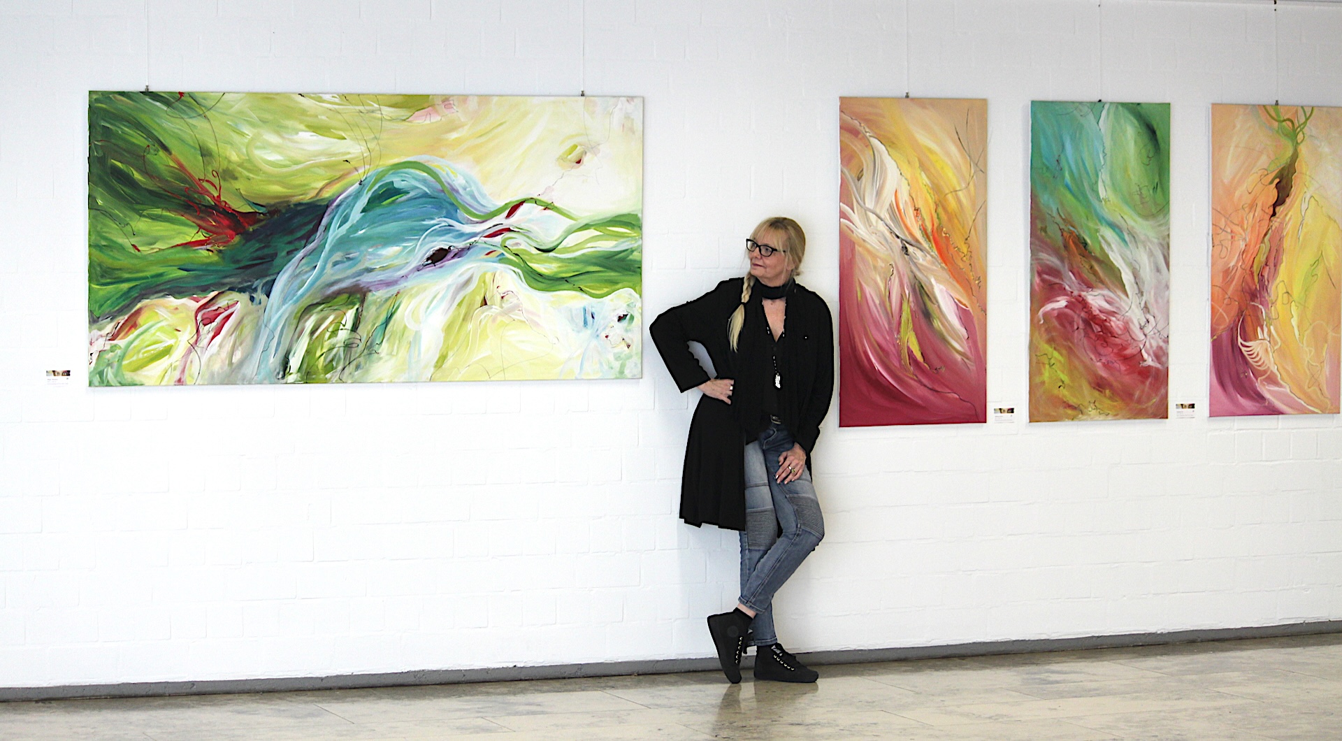 Silvia Grasser lehnt in einer Ausstellung an der Wand zwischen ihren großformatigen, farbenfrohen Acrylbildern