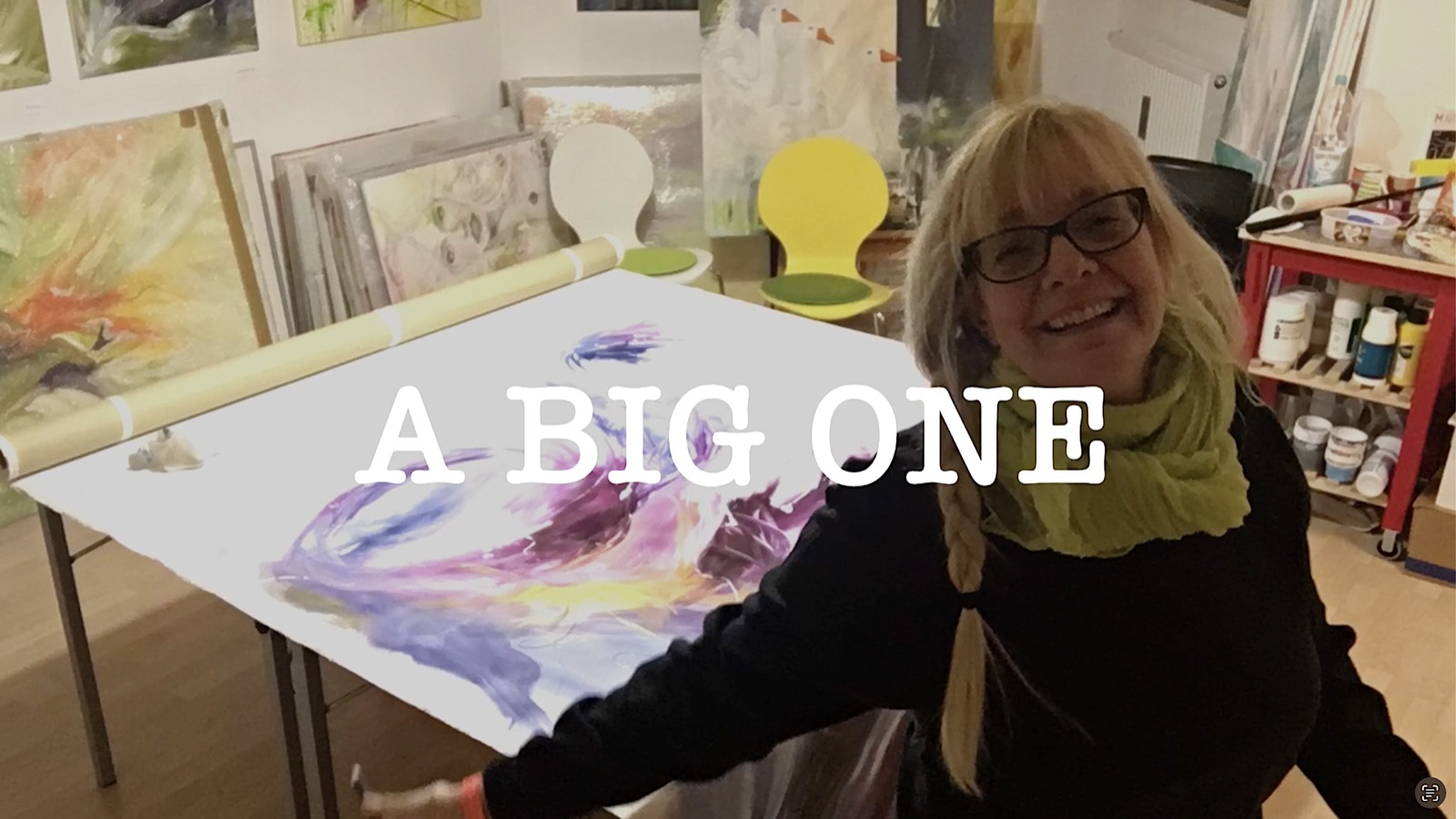 Silvia Grasser lacht im Atelier neben einem großen Bild in Violett und Gelb, das auf dem Tisch liegt